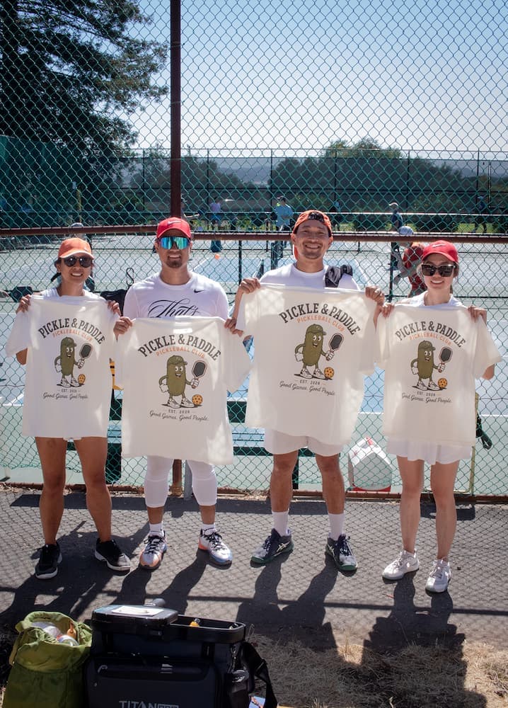 Four players holding up Pickle & Paddle club t-shirts courtside